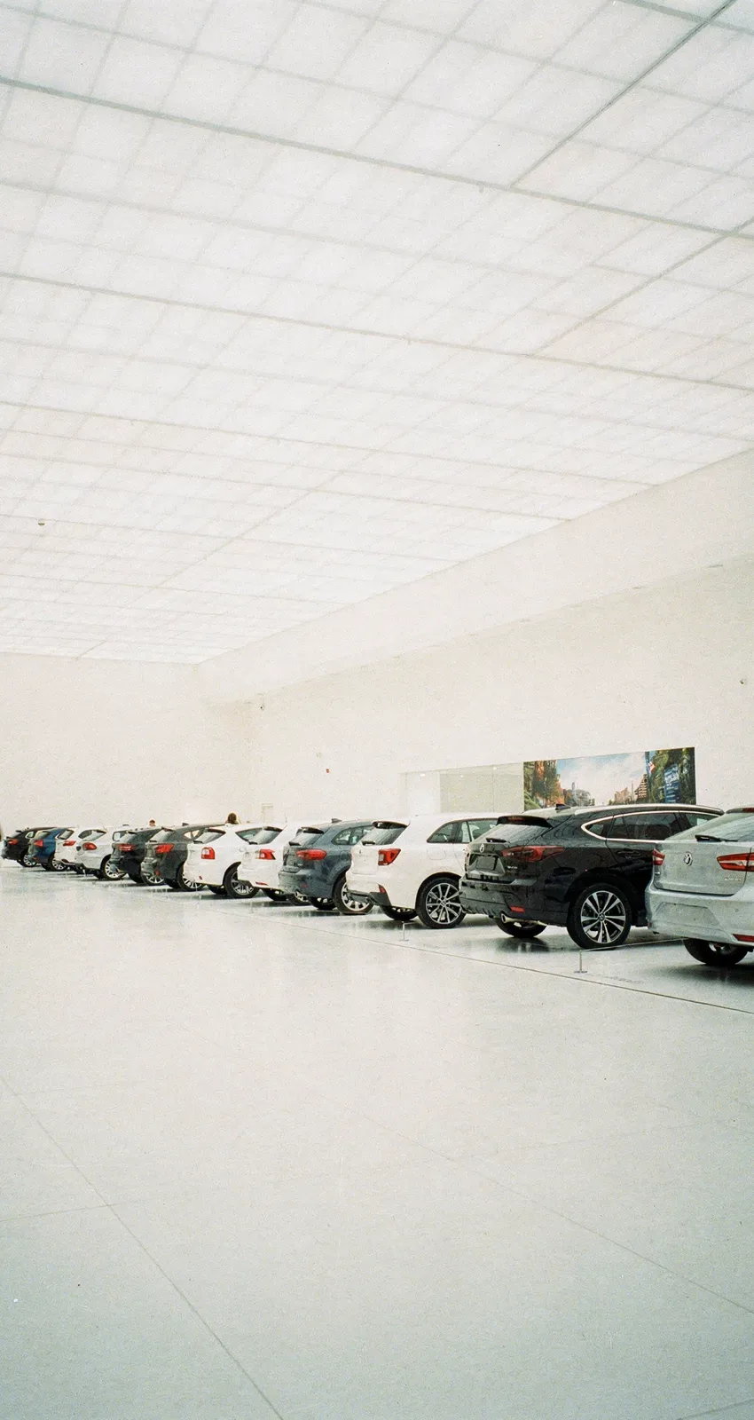 Sehr helle Ausstellungshalle mit Lichtdecke, eine Reihe Fahrzeuge von hinten, eine Person geht bewegungsunscharf durchs Bild, KI-generiert