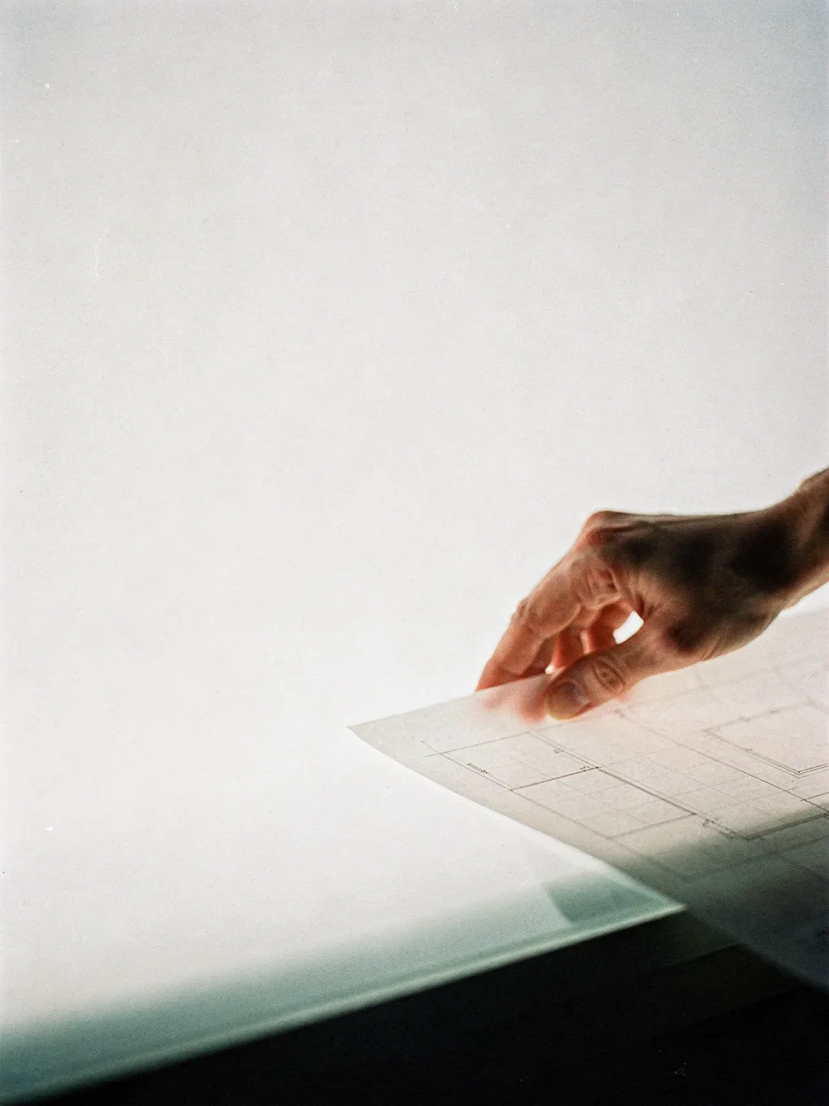Eine Hand legt ein Transparentpapier mit Layoutraster auf einen hell durchleuchteten Zeichentisch, daneben eine große helle Leerfläche, KI-generiert