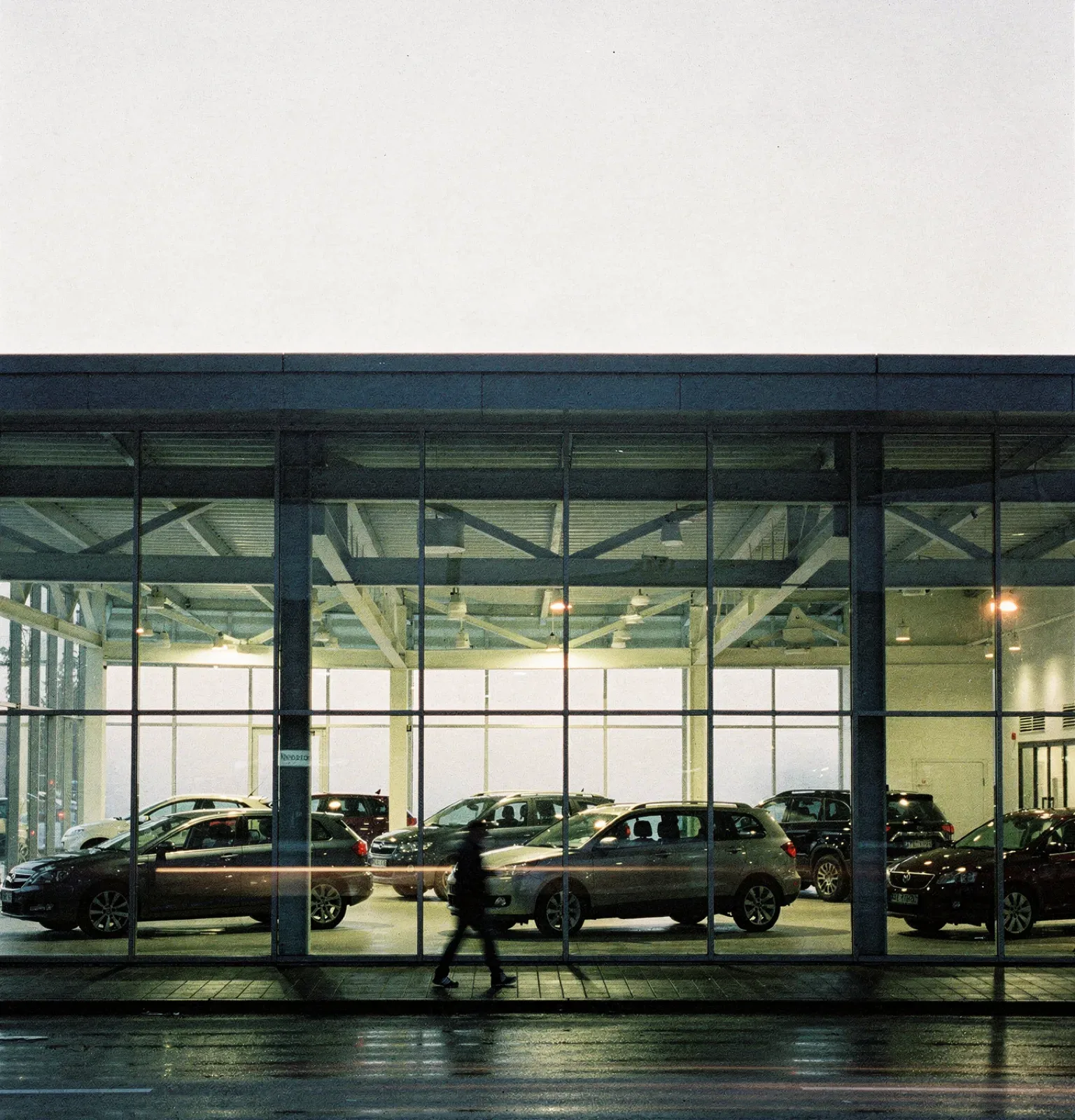 Autohaus hinter einer hohen Glasfront im Gegenlicht, Fahrzeuge als dunkle Silhouetten, eine Person geht bewegungsunscharf vorbei, KI-generiert