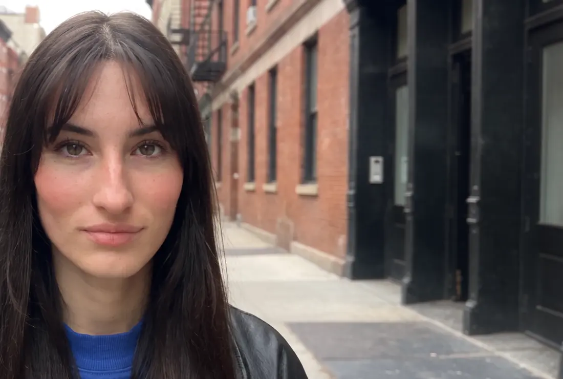Lune, eine junge Frau mit dunklem Pony und Sommersprossen, steht im Tageslicht auf einem Gehweg in New York, schaut in die Kamera und lacht los, KI-generiert